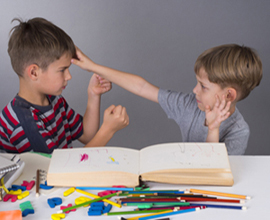 Dos niños sentados a la mesa, uno con los puños apretados y el otro con la mano en la frente del primer niño.