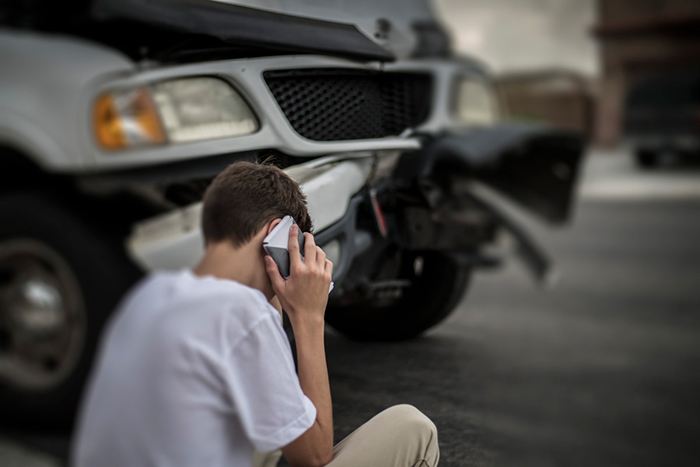Imagen de un adolescente sentado en la acera hablando por celular frente a un auto con daños por colisión frontal.