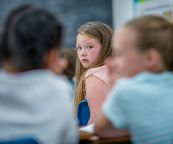 Una niña está sentada en una sala de clases, mirando por sobre su hombro a dos compañeras de curso que están cuchicheando entre ellas.